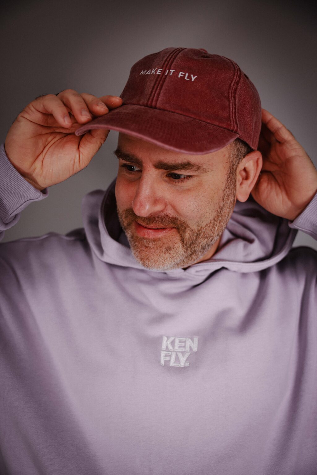 Oliver Margrander lächelt nach unten und hält mit beiden Händen seine Kappe, dazu trägt er einen Hoodie mit KEN-FLY-Logo