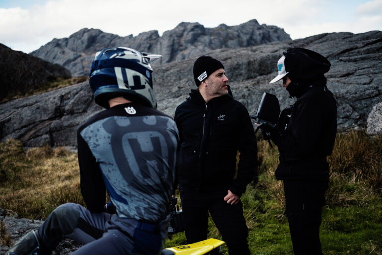 Behind the scenes: Ein Fahrer in Motocross-Ausrüstung mit Helm und zwei Crewmitglieder in schwarzer Winterkleidung stehen zwischen Felsen.
