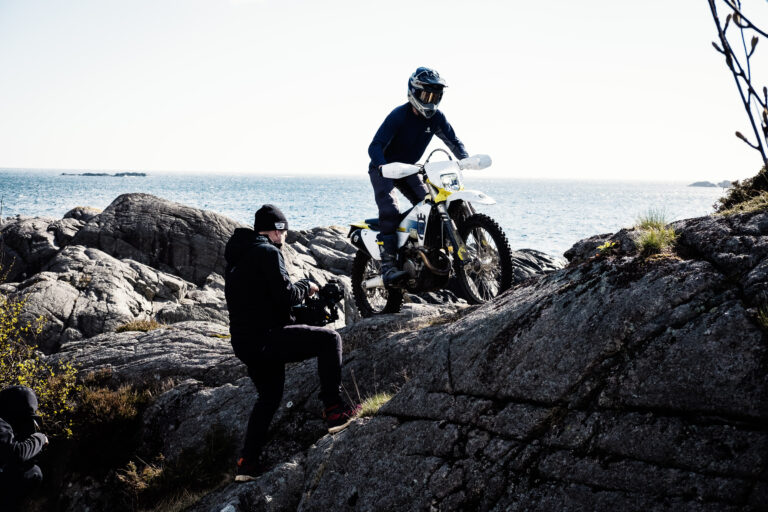 Behind the scenes: Ein Fahrer auf einem Offroad-Motorrad steht auf Felsen am Meer, links daneben arbeitet ein Crewmitglied.