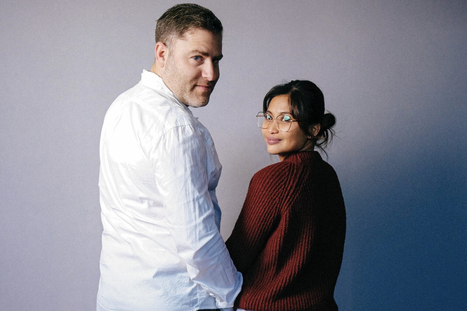 Oli und Rigene, die Gastgeber des Podcasts „Is it?“, stehen Seite an Seite und blicken über die Schulter in die Kamera.