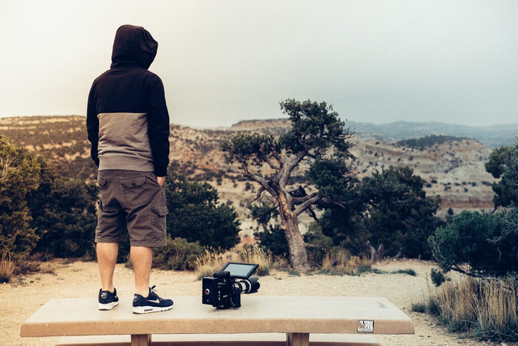 Filmemacher mit Kamera auf einer Steinbank vor weiter Berglandschaft